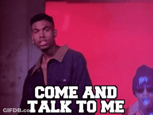 A Man In A Denim Jacket Is Standing In Front Of A Red Wall And Talking To Another Man . GIF