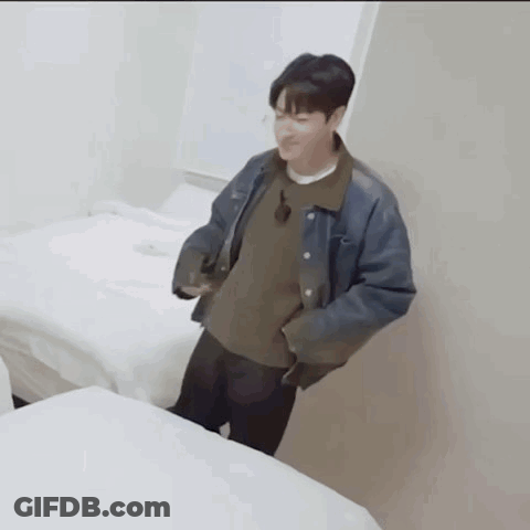 A Man In A Denim Jacket Is Standing Next To A Bed In A Room GIF