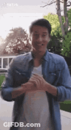 A Man In A Denim Shirt And White Shirt Is Standing In Front Of A Tree . GIF