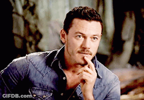 A Man In A Denim Shirt Holds His Hand To His Chin And Looks At The Camera GIF