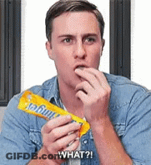 A Man In A Denim Shirt Is Eating A Candy Bar And Saying What ? GIF