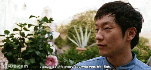 A Man In A Denim Shirt Is Talking To Another Man In Front Of A Garden . GIF