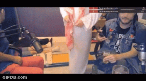 A Man In A Denim Vest Is Sitting In Front Of A Microphone While A Woman Stands In Front Of Him GIF