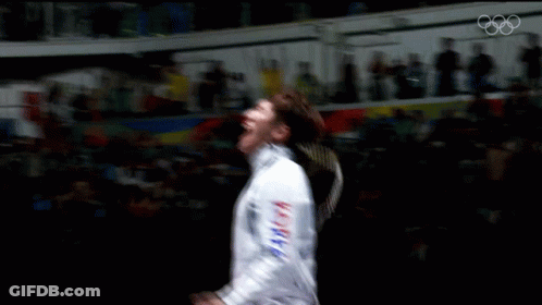 A Man In A Fencing Uniform Is Dancing In Front Of A Crowd With The Olympics Logo In The Background GIF