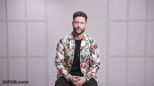 A Man In A Floral Jacket Is Sitting In Front Of A Wall With The Words Love Conquers All Above Him GIF