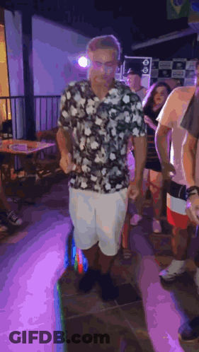A Man In A Floral Shirt And White Shorts Is Dancing In A Crowd Of People At A Party . GIF