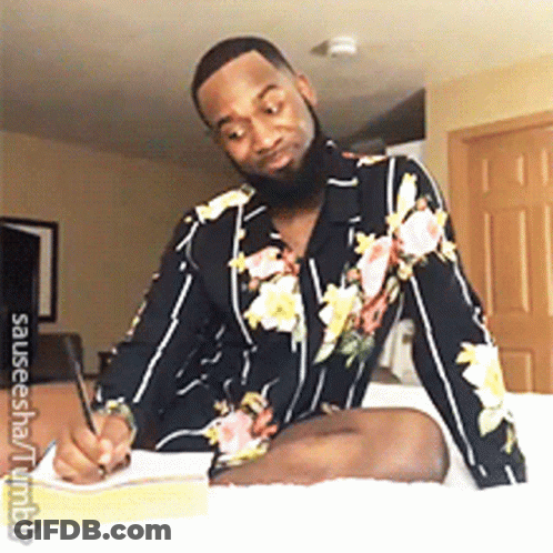 A Man In A Floral Shirt Is Sitting On A Bed Writing In A Notebook GIF