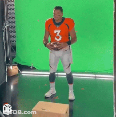 A Man In A Football Uniform Is Standing On A Box Holding A Football In Front Of A Green Screen . GIF