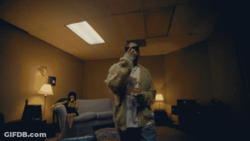 A Man In A Fur Coat Stands In A Dark Room With A Woman Sitting On A Couch GIF