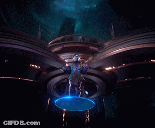 A Man In A Futuristic Outfit Is Standing In A Dark Room With A Blue Light Behind Him GIF