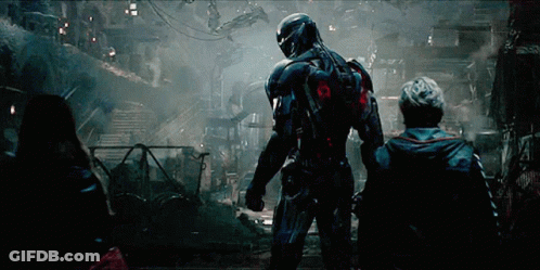 A Man In A Futuristic Suit Stands In A Dark Room With Two Other People GIF