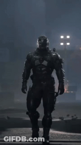 A Man In A Futuristic Suit With A Helmet On Stands In The Dark GIF