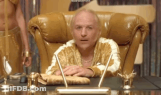 A Man In A Gold Robe Is Sitting At A Desk With A Pen . GIF