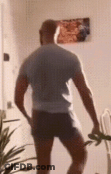 A Man In A Gray Shirt And Black Shorts Is Standing In A Room With A Picture On The Wall Behind Him . GIF