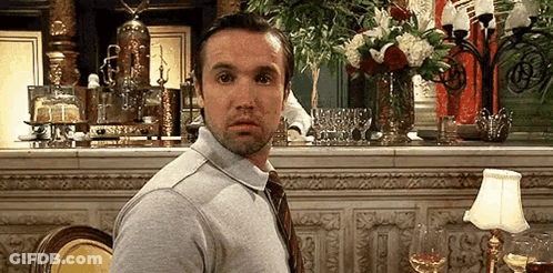 A Man In A Gray Shirt And Tie Is Sitting At A Table In A Restaurant With A Vase Of Flowers In The Background . GIF