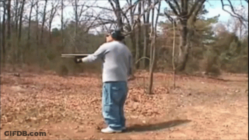 A Man In A Gray Shirt Is Holding A Shotgun In A Field GIF