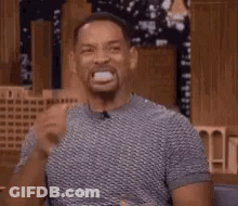 A Man In A Gray Shirt Is Making A Funny Face While Sitting At A Table . GIF
