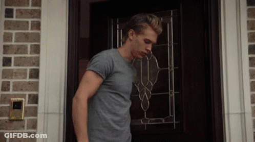 A Man In A Gray Shirt Is Standing In Front Of A Door GIF