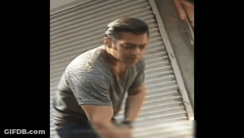 A Man In A Gray Shirt Is Standing In Front Of A Shutter GIF