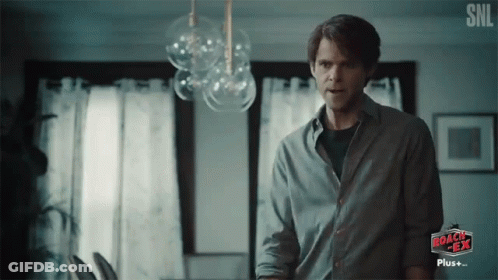 A Man In A Gray Shirt Is Standing In Front Of A Sign That Says Roach Tea GIF