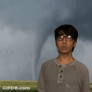 A Man In A Gray Shirt Is Standing In Front Of A Tornado GIF
