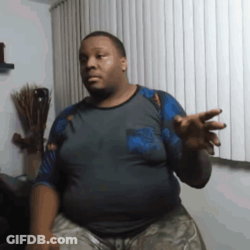A Man In A Gray Shirt Is Standing In Front Of A Window With Blinds GIF