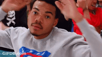 A Man In A Gray Sweatshirt Is Making A Funny Face While Sitting In A Crowd Of People . GIF