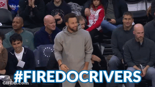 A Man In A Gray Sweatshirt Stands In Front Of A Crowd With The Hashtag #Firedocrivers Above Him GIF