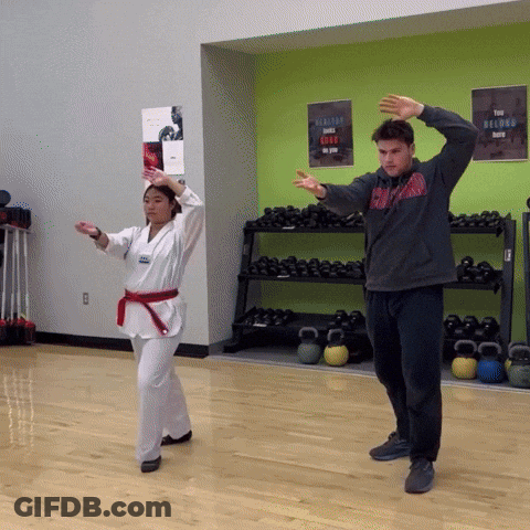 A Man In A Gray Sweatshirt Stands Next To A Woman In A Karate Uniform GIF
