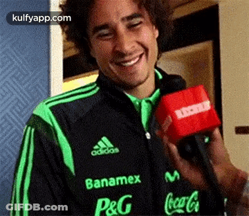 A Man In A Green And Black Adidas Jacket Is Holding A Red Microphone And Smiling . GIF