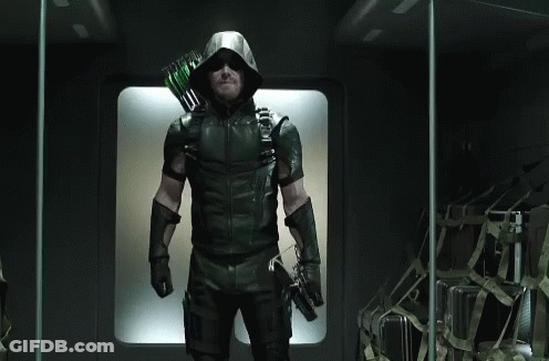 A Man In A Green Arrow Costume Is Standing In A Dark Room Holding A Bow And Arrow . GIF