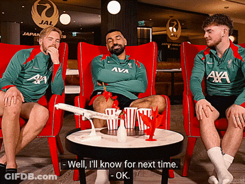 A Man In A Green Aya Shirt Sits With Two Other Men GIF