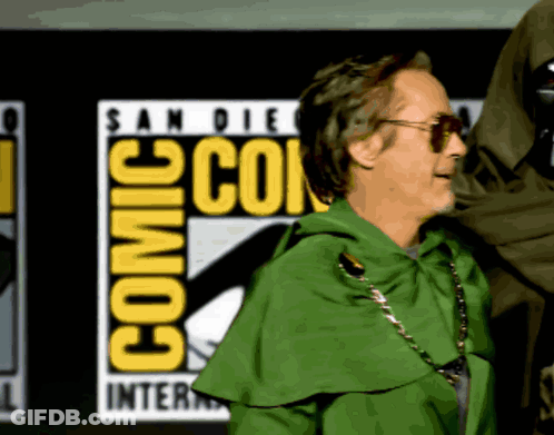 A Man In A Green Cape Is Standing In Front Of A Comic Con Logo GIF