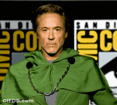 A Man In A Green Cape Stands In Front Of A Comic Con Sign GIF