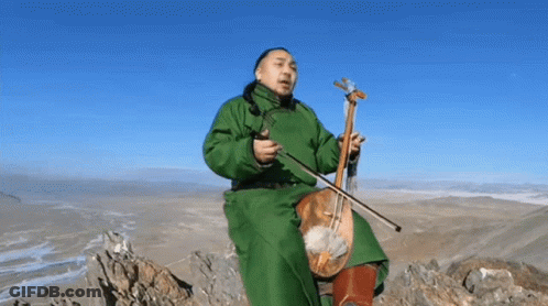 A Man In A Green Coat Is Sitting On Top Of A Mountain Playing A Violin . GIF