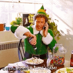 A Man In A Green Elf Costume Is Talking On A Cell Phone While Pouring Popcorn Into A Bowl . GIF