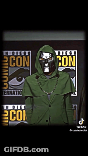 A Man In A Green Hooded Jacket Stands In Front Of A Comic Con Poster GIF