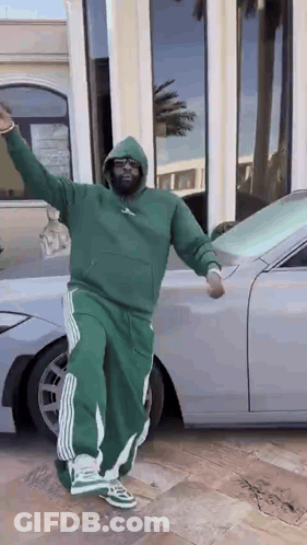 A Man In A Green Hoodie And Green Pants Is Standing In Front Of A Silver Car . GIF