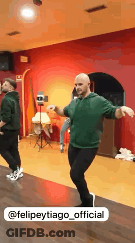 A Man In A Green Hoodie Is Dancing In A Room With A Red Wall Behind Him GIF