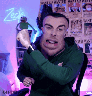 A Man In A Green Hoodie Is Sitting In Front Of Wanted Posters GIF
