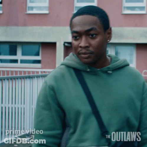 A Man In A Green Hoodie Is Standing In Front Of A Building With The Outlaws Written On It GIF