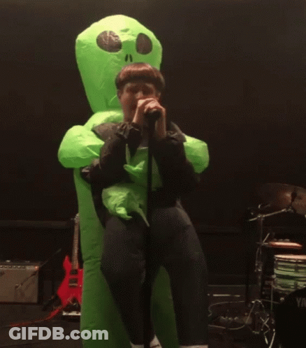 A Man In A Green Inflatable Alien Costume Singing Into A Microphone GIF