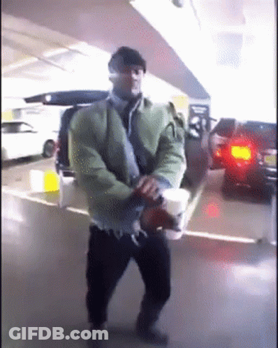 A Man In A Green Jacket Is Holding A Cup Of Coffee In A Parking Garage GIF