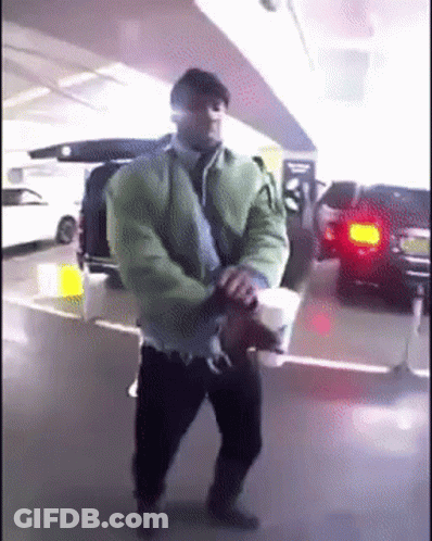 A Man In A Green Jacket Is Holding A Cup Of Coffee In A Parking Garage GIF