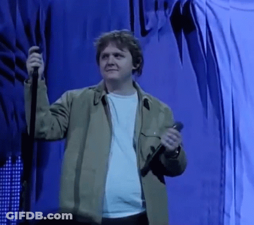 A Man In A Green Jacket Is Holding A Microphone On A Stage In Front Of A Blue Curtain . GIF