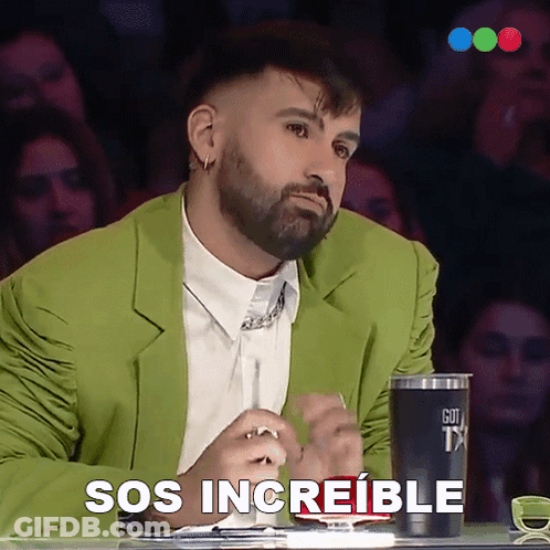 A Man In A Green Jacket Is Sitting At A Table With A Cup That Says Sos Increible On It GIF