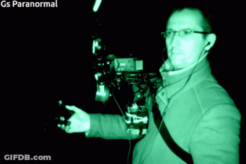 A Man In A Green Jacket Is Standing In A Dark Room With Gs Paranormal Written On The Bottom Right GIF