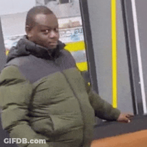 A Man In A Green Jacket Is Standing In Front Of A Door And Looking At The Camera . GIF