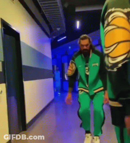 A Man In A Green Jacket Is Walking Down A Hallway With Another Man . GIF