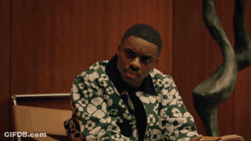 A Man In A Green Jacket Sits In A Chair In Front Of A Wooden Wall GIF